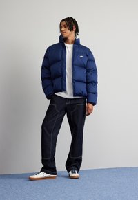 Ναυτικό φουσκωτό μπουφάν με ψηλό γιακά και μπροστινό φερμουάρ, φορεμένο πάνω από ένα λευκό μπλουζάκι, σε συνδυασμό με σκούρα τζιν και λευκά αθλητικά παπούτσια.