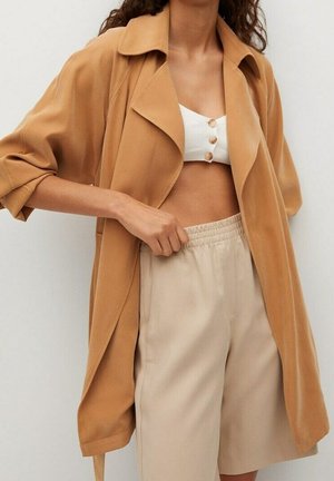 Femme portant un trench-coat ouvert beige sur un crop top blanc boutonné et un short beige à taille élastique, debout, ajustant son short d’une main.
