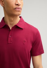 Weinrotes Polohemd aus glattem Baumwollstoff. Mit einem Zwei-Knopf-Verschluss und einem kleinen, gestickten Logo auf der linken Brust. Kurze Ärmel.