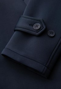 Tejido azul oscuro de un puño de prenda que presenta un diseño elegante, dobladillo doblado y dos botones circulares negros con una superficie texturada.