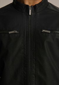 Czarna kurtka z sztucznej skóry z wysokim kołnierzem, dwoma kieszeniami na zamek na klatce piersiowej oraz teksturowaną powierzchnią z gładkimi akcentami. Elementy zamka są metalowe.