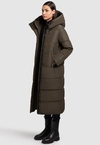 Ilgas paminkštintas paltas tamsiai alyvuogių žalios spalvos, su aukštu apykakliu, didele kapišonu, užtrauktuku uždarymu ir šoninėmis kišenėmis. Derinamas su juodais aulinukais.