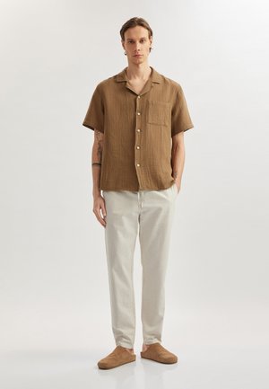 Brown short-sleeve, textured button-up shirt with a chest pocket, paired with light-colored trousers and tan slip-on shoes.