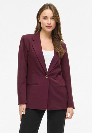 Nő bordó egysoros gombos blézerben, fehér felsővel és fekete nadrággal, világos egyszínű háttér előtt állva.