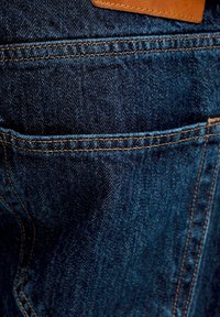 Donkere blauwe spijkerbroekzak met zichtbare oranje stiksels en een kleine bruine leren patch boven de zak.