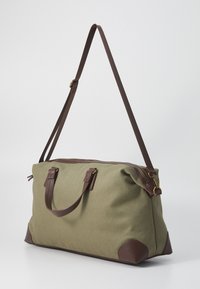 Grønn stoff totebag med brune lærdetaljer, glidelås, to korte håndtak og en lang justerbar stropp. Teksturert overflate med rektangulær form.