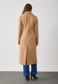 Lang beige jas met een rechte silhoutte, brede kraag en achterventilatie, gecombineerd met blauwe flare jeans en zwarte hoge hakken.