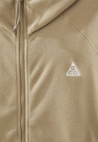 Beige Fleece-Zip-Jacke mit einem dreieckigen weißen Logo auf der linken Brust, glatter Textur und verstellbarem Kapuzenzug.