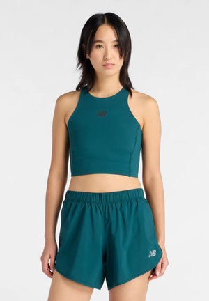 Femme portant un crop top sans manches turquoise et un short de sport assorti, debout devant un fond blanc.
