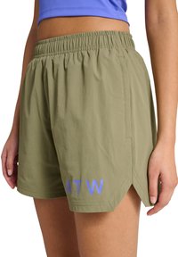 Frau trägt olivgrüne sportliche Shorts mit elastischem Bund und blauen "ATW"-Buchstaben nahe dem Saum, kombiniert mit einem lila bauchfreien Oberteil.