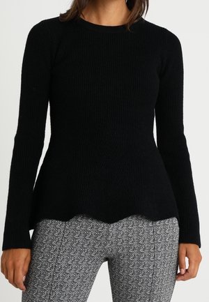 Haut noir à manches longues en côte avec une silhouette peplum, tissu texturé et ourlet en dents de scie, associé à un pantalon gris à motifs.
