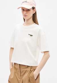 Camiseta blanca de algodón con mangas cortas y cuello redondo, con bordado verde de "Lacoste". El modelo lleva una gorra rosa claro.
