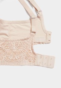 Reggiseno beige chiaro con dettagli in pizzo, spalline regolabili e ganci per la chiusura. Texture liscia con motivo floreale in pizzo su un lato.