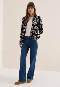 Cardigan noir et crème à imprimé léopard porté sur une chemise blanche, associé à un jean large bleu et des baskets marron. Texture lisse, design décontracté.