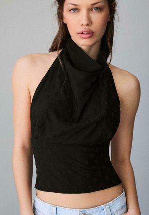 Ung kvinde med blå øjne iført en sort, ærmeløs haltertop og lyseblå jeans, stående foran en grå baggrund.