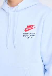 Šviesiai mėlynas „Nike“ džemperis su priekiniais kišenėmis. Yra raudonas logotipas ir juodas tekstas „TIK ĮGALIOTAM PERSONNEL“. Minkšta medžiaga, reguliuojama kapuča.