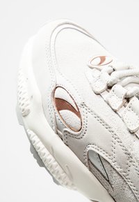 Weiße Sneaker mit einer Kombination aus glattem und strukturiertem Leder, runder Form, gepolsterter Zunge, aufwendigen Nähten und einem bronzefarbenen Akzent.
