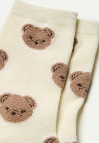 Knæhøje sokker i creme med brune teddybjørnsmønstre. Har blød frottéstruktur og glat ribbede top. Materiale af bomuldsblanding.