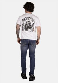 T-shirt bianca in cotone con una stampa grafica nera di un leone ruggente e la parola "PROPAGANDA" sul retro. Indossata con jeans blu.