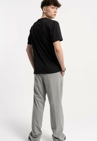 Młody mężczyzna stoi z rękami w kieszeniach, ubrany w czarną koszulkę i jasnoszare spodnie w prążki, patrząc przez ramię.