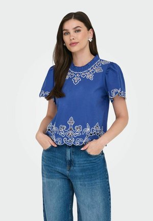 Blouse bleue à manches courtes avec des motifs floraux brodés blancs, un col froncé et un ourlet légèrement court, associée à un jean bleu.