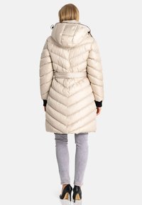 Beige Daunenjacke mit einem Zickzackmuster, Kapuze und Kordelzug. Mit einem Gürtel und schwarzen Handgelenkbündchen ausgestattet. Getragen über grauen Jeans und hohen Schuhen.
