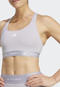 Lichtgrijze sportbeha met een soepele textuur, brede elastische band met "TECHFIT" branding en een minimalistisch ontwerp met subtiele logo-accenten.