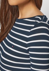 Femme portant une chemise rayée horizontale bleu marine et blanche avec un petit logo rouge, blanc et bleu sur la poitrine, et de longs cheveux bruns visibles.