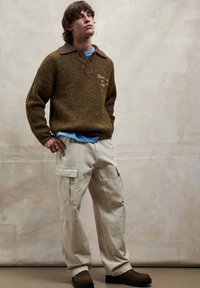 Ruskea ribbineulepaita, jossa tumma kaulus, vaaleansininen t-paita alla ja beige farkkutyyppiset housut; malli, jolla on ruskeat kengät.