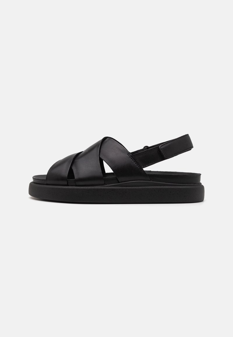Sandale en cuir noir avec larges brides croisées, bride arrière ajustable et semelle épaisse rembourrée, présentée de profil sur un fond uni.