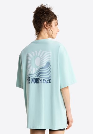 Woman with curly hair wearing light blue oversized T-shirt with sun and wave graphic and "THE NORTH FACE" text on the back.