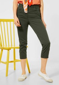 Kvinna klädd i olivgröna croppade byxor, vita slip-on skor, orange topp med matchande scarf, står bredvid en gul trästol.