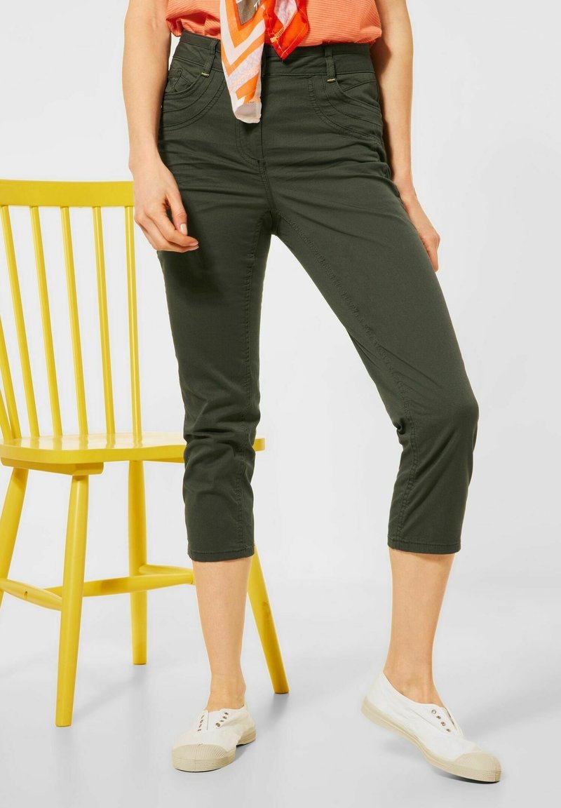 Kvinna klädd i olivgröna croppade byxor, vita slip-on skor, orange topp med matchande scarf, står bredvid en gul trästol.