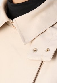 Gros plan d'un col beige avec deux boutons-pression métalliques sur un vêtement à col roulé noir.