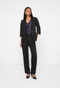 Žena stojí a má na sebe čierne sako s ¾ rukávmi, čierne široké nohavice, čierne lodičky a fialový top s flitrami, na bielom pozadí.