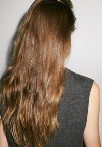 Lang, golvend bruin haar valt over de rug en contrasteert met een grijze mouwloze gebreide top met een textuurafwerking.