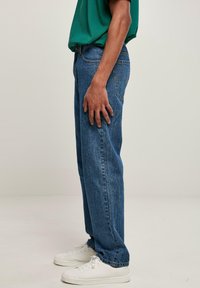 Vaqueros de mezclilla azul claro con corte recto, que cuentan con el clásico diseño de cinco bolsillos, usados con zapatillas blancas y una camisa verde.