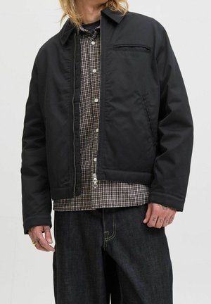 Mand iført sort lynlåstrøje over en brun og sort ternet skjorte med mørke jeans, viser tatoveringer på fingrene.