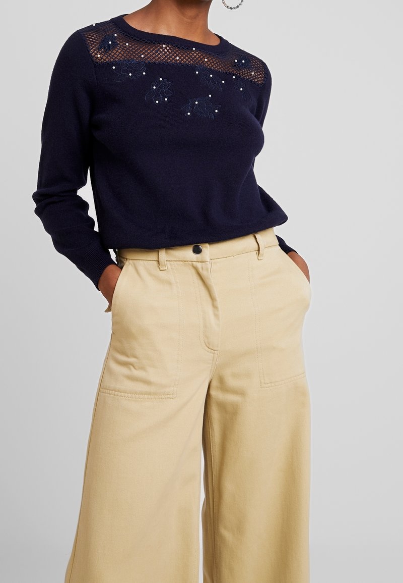 Pull à manches longues bleu marine avec broderie florale et empiècement en maille, associé à un pantalon beige taille haute à jambes larges avec poches avant.