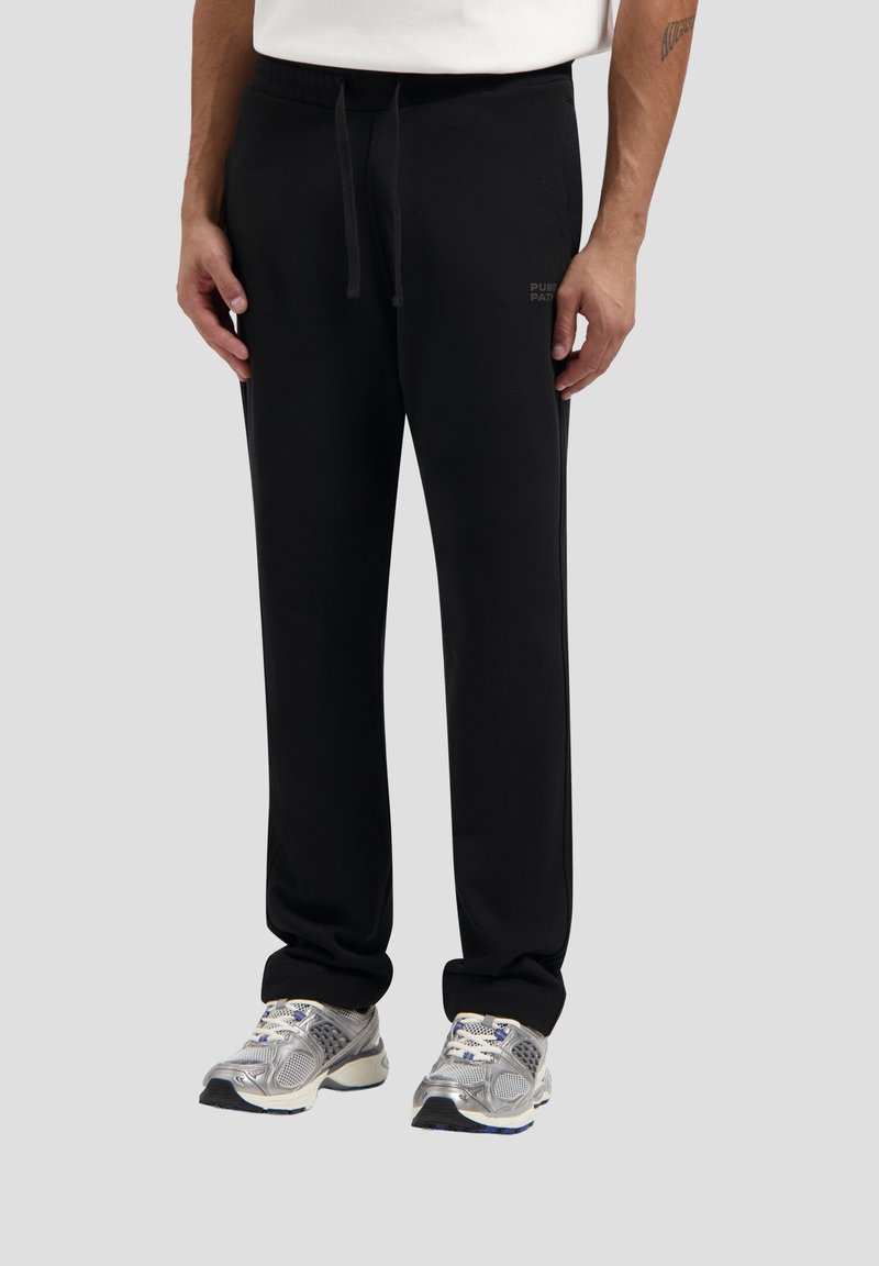 Schwarze Sweatpants mit elastischem Bund und Kordelzug. Sie verfügen über seitliche Taschen und dezente Branding-Elemente. Kombiniert mit silbernen Sportschuhen.