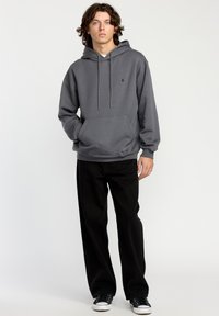 Grå hættebluse med en frontlomme, snørebånd og et lille logo. Bæres med sorte bukser og sorte sneakers, mod en ensfarvet baggrund.