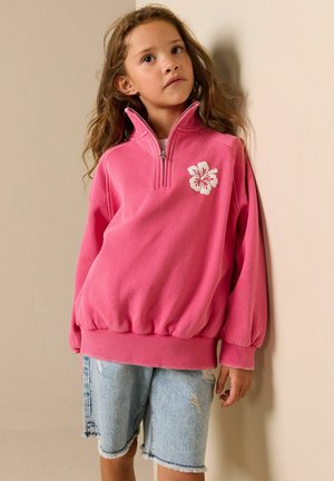 Sweat-shirt à zip rose avec un motif floral sur la poitrine, doté d'une coupe décontractée et d'un ourlet côtelé, associé à un short en jean déchiré.