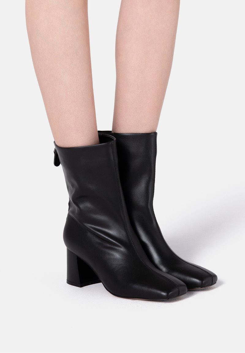 Bottes noires en cuir à la cheville avec fermeture éclair, talon bloc, bout carré et texture lisse. Détails cousus le long des coutures.