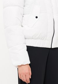 Witte pufferjas met ritsluiting, elastische zoom en een flapzak met zwarte knopen. Stevige textuur, cropped ontwerp en brede mouwen.
