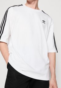 Bílý bavlněný tričko s černým detailním třípruhovým vzorem na ramenou a malým černým logem Adidas na hrudi. Krátké rukávy, volný střih.