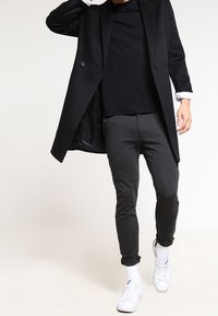 Cappotto doppiopetto nero sopra una camicia nera, abbinato a pantaloni slim-fit grigio scuro e sneakers bianche. Texture lisce e linee pulite.
