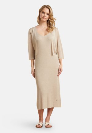 Beige gebreide lange jurk met een ronde halslijn, driekwartmouwen en een aansluitend lijfje dat uitloopt in een wijd uitlopende zoom. Vergezeld van een cropped cardigan.