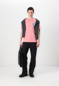 Lyse rosa T-skjorte med en blå og grønn grafisk lomme, sammen med svarte jeans, svarte sko og en svart totebag. Grå hettegenser slengt over skuldrene.