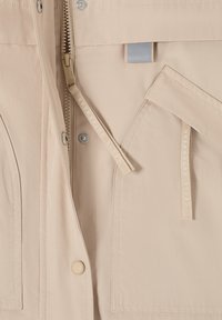 Veste beige avec fermeture éclair, boutons-pression, poches à rabat et languettes en tissu portant l'inscription "CECIL OUTERWEAR".