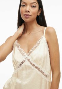 Kremfarget satengkamisole med delikat blonderkant langs V-uttak og skuldre. Har justerbare spaghettistropper og mykt, flytende stoff.